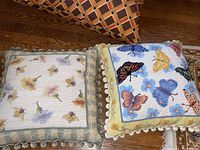 Two needlepoint pillows on floor: one with butterfly design and pom-pom border, one with bee and flower design with green checked border and tassel trim, plus corner of geometric fabric pillow visible