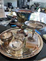 Full view of the 9-piece silverplate collection arranged on a black countertop. Visible are the large engraved serving platter, champagne bucket, candy dish, four small bowls, wine charger, boxed toothpicks, and a trivet.