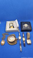 Group view of pendant watch, pocket watch, three wristwatches and two desk clocks on blue background