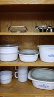 Front view of wooden shelves with multiple CorningWare baking dishes and ramekins stacked, showing lids and blue floral patterns.
