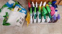 Wide shot of bin with multiple spray bottles of cleaning products with various colored nozzles on the floor next to a pack of paper towels
