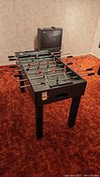 Foosball table fully set up in room with red carpet and beige walls. Shows multiple player rods with handles and players in red and blue colors.