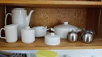 Full view of white porcelain tea service pieces and two stainless steel grinders on shelf