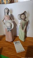Pair of two high-gloss Lladro porcelain figurines of girls on display, one in pink dress holding a hat, the other in green dress wearing a hat