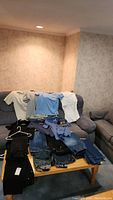 Overview photo of men's clothing including 6 shirts and multiple pairs of jeans laid out on a table and couch, showing clothing condition and variety