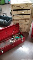 Cream metal cabinet with multiple drawers, visible scratches, rust, and handwriting on drawer fronts. Adjacent red metal toolbox on floor.