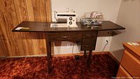 Sewing machine mounted in wood table with accessories on top and drawers beneath, viewed from front.