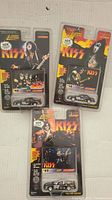 Photo of 3 Johnny Lightning KISS diecast car packages against wall, showing front of packaging with band member images and car models visible