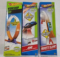 Front view of three boxed Hot Wheels track sets: Workshop Loop, Jump, and Gravity Clamp, showing track piece count and colorful packaging.
