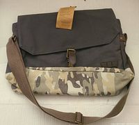 Front view of the Levi's camo messenger bag showing dark gray upper fabric, camo print lower, leather strap with metal buckle, and adjustable brown shoulder strap.