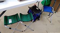Two green folding stools with metal tubular legs and fabric seats beside a camping table with blue storage bag underneath.