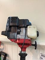Side view showing the gas trimmer engine with pull start and vented black cover, red body, and white fuel tank.