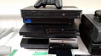 All four PlayStation consoles stacked: PS2 units, PS3, and PS1 visible with one controller on top.