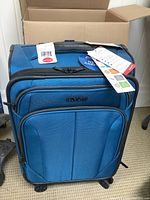Front view of blue American Tourister suitcase in box, tags visible including brand and product line.