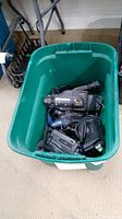 Green plastic bin containing five vintage handheld video cameras from Sony, GE, Canon, Sanyo, and Zeeland.