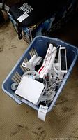 Blue basket containing two white Nintendo Wii consoles and several white Wii remote controllers along with steering wheel and hockey stick attachments scattered in the basket.