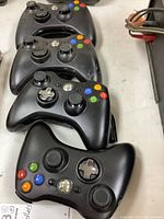Four black Xbox 360 wireless controllers laid out in a row showing top/down view with all buttons and analog sticks visible.