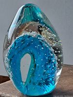 Front angled view of clear glass egg-shaped paperweight showing embedded blue wave pattern with bubbles inside.