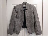 Front view of gray textured women's blazer hanging on hanger against white door, showing open collarless design and asymmetrical hem