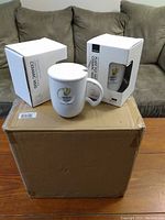 Photo showing two ceramic mugs in individual white boxes, one mug with handle facing forward and the other visible behind the box, all placed on large cardboard box.