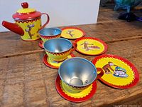 Photo showing teapot, tea cups, saucers, and plates with Curious George graphics, red and yellow color scheme on wooden floor.