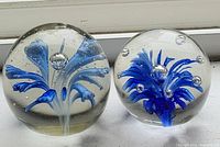 Two glass paperweights featuring intricate blue flower designs encased in clear glass, positioned on a white surface near a window.