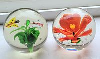 Photo of two spherical glass paperweights, one with an orange flower and one with a green plant and two butterflies inside.