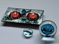 Photo showing glass tea light holder with hummingbird design holding two lit tea light candles, next to a spherical blue swirl glass paperweight and small glass heart