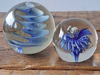 Photo showing front view of two round clear glass paperweights on wooden surface; larger with blue swirl inside, smaller with blue flower-like image and bubbles.