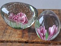 Photo showing two glass paperweights with pink flower images; one oblong with bubbles and detailed flower, the other rounded with petals