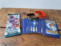 Photo of unopened Japanese Pokemon card pack, over 50 loose cards showing their backs, and two Pokemon-themed silicone bracelets on a wooden surface.