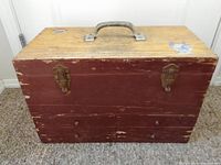 Front view of wooden storage box painted red with metal handle and two metal latch clasps, visible wear on paint.