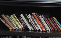 A photo showing a row of 18 books covering various topics such as cooking, music, history, and biographies, mostly hardcover and some paperbacks.