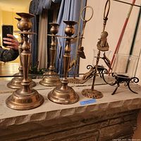 Three brass candle stick holders arranged side by side with a decorative note holder shaped like a hand holding a ring on stand, placed on a stone ledge in front of a mirror.