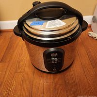 Closed view of the stainless steel electric pressure cooker with control panel and black handle on wood floor.