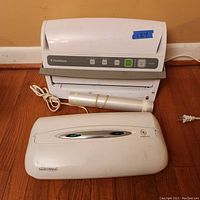Front view of FoodSaver V3240 vacuum sealing system, with roll of sealing bags and Rival Seal-a-Meal unit on the floor in front.