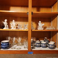 Shelf with four white angel figurines, two clear star-shaped candle holders, blue and white polka dot pottery cups, and assorted clear glass drinkware.