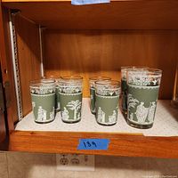 Set of six green tumblers featuring detailed white classical Hellenic scenes and decorative borders, displayed on a wooden shelf.
