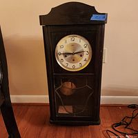 Front view of large solid wood wall clock with white dial, black hands and numerals, and glass door covering the pendulum area.