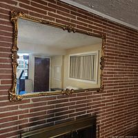 Full view of large rectangular vintage mirror with ornate gold framing hanging on a brick wall, showing size and setting.
