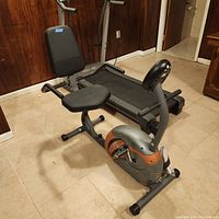 Side view of the MPEX Marcy recumbent bike showing padded seat with backrest, frame, pedals, and part of treadmill in the background.