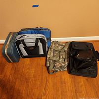 Photo of five assorted travel bags standing on wooden floor against peach wall, showing a variety of styles and colors including dark blue hard case, gray-black soft bag, camo backpack, blue-gray bag, and black rolling bag.