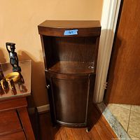 Wooden corner cabinet with a dark finish, curved front door with knob, and open shelf above it, placed against a wall near a door and other wooden furniture.