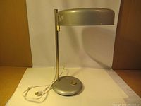 Full view of lamp showing curved shade, vertical metal stem, round base with switch and attached power cord with plug.