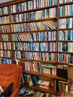Photo showing multiple wooden shelves filled with books, some leaning and stacked horizontally, with some boxes of books at the bottom.