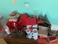 Photo of assorted Christmas decorations on table including red glass candle holders, boxed ornaments, mugs, greenery, and ceramic tree figurine