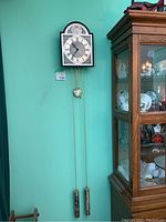 Full view of Temporis Campus Fugit wall clock hanging on teal wall next to wooden display cabinet, showing two brass weights and pendulum on metal chains.