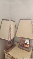 Pair of metal table lamps with square bases and beige tapered lampshades featuring copper embossed floral trim