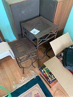 Pair of metal plant stands positioned on a wooden floor next to a corner screen. Both stands have square tops with perforated surface. Side view showing lattice metal design and curved three-legged supports with circular bases.