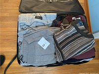 Top view of multiple thick knitted sweaters and sweater vests in neutral and earth tones inside a partially packed suitcase on wooden floor.